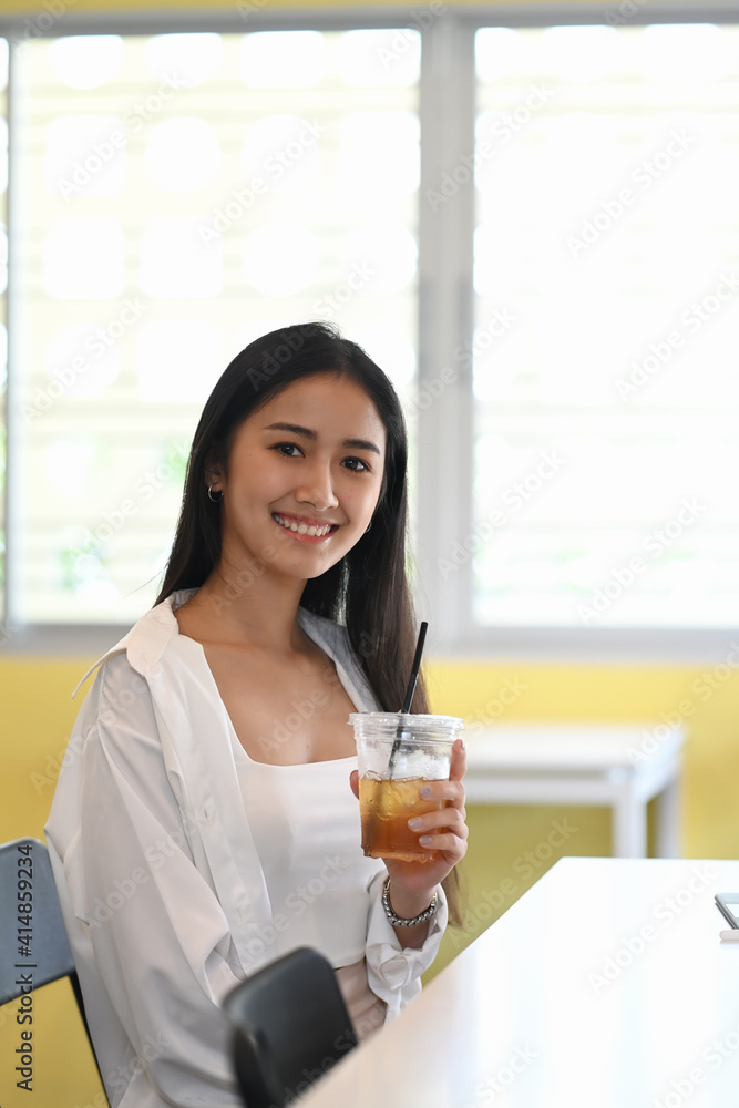 Portrait of cheerful young woman holding ice coffee and smiling to camera while sitting in office. Portrait of cheerful young woman holding ice coffee and smiling to camera while sitting in office.