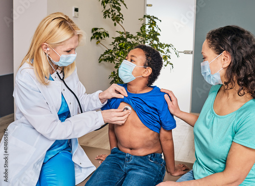 Pediatrician auscultating boy's lungs using a stethoscope. African American child has pneumonia or coronavirus, near mother wearing protective mask photo