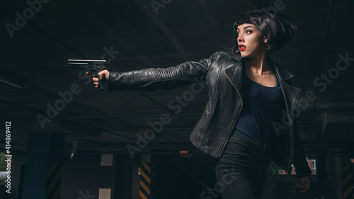 Beautiful brunette sexy spy agent (killer or police) woman in leather jacket and jeans with a gun in her hand running after someone, to catch him on parking