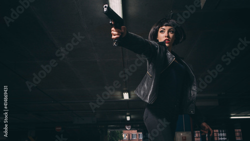 Beautiful brunette sexy spy agent (killer or police) woman in leather jacket and jeans with a gun in her hand running after someone, to catch him on parking