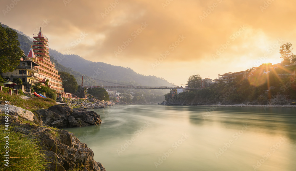 Stunning view of the Ganga river embankment with the Lakshman Jhula bridge and Tera Manzil Temple (Trimbakeshwar) during a beautiful sunset. Rishikesh, Uttarakhand, India.