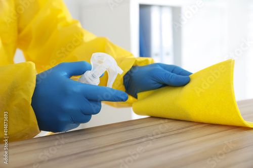 Employee in protective suit and gloves sanitizing wooden countertop indoors, closeup. Medical disinfection