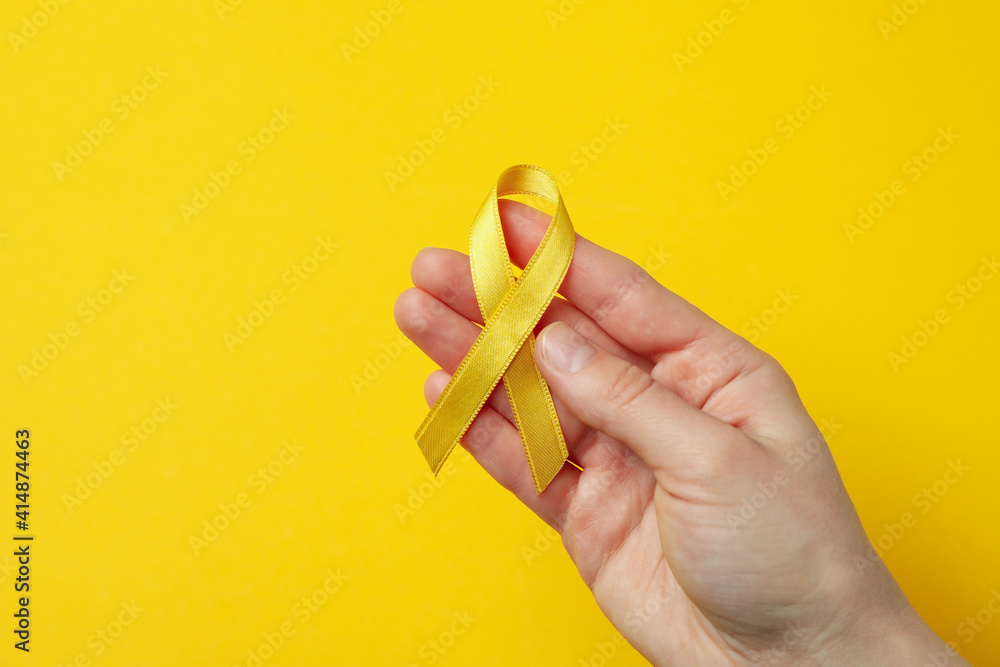 Female hand hold child cancer awareness ribbon on yellow background Female hand hold child cancer awareness ribbon on yellow background
