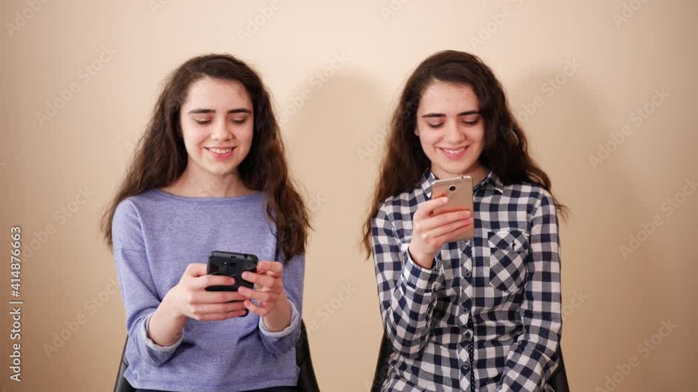 Two teenager twin sisters girls stay at home with mobile smartphones have fun sitting on spinning chairs