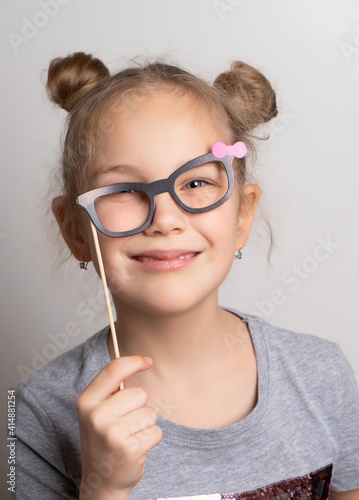 Fototapeta Naklejka Na Ścianę i Meble -  cheerful crazy little girl looks through rickety paper glasses. Festive costume for a masquerade. Attractive female child posing with photo booth accessory studio portrait shot