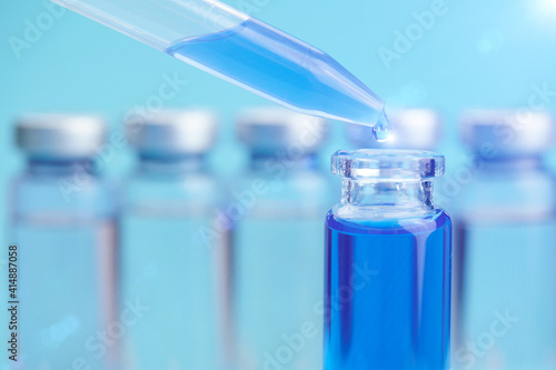 Pipette with liquid and bottle. The vaccine against the coronavirus is blue. A drop of liquid flows out of the pipette and is shot in close-up. A glint of light on a drop. Blue background.