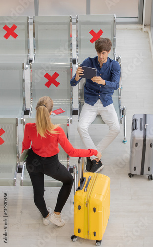 Male tourist sitting on row seats in airport lobby chair with social distancing sign and using tablet computer to take photo of female traveler who comes with yellow language. New normal journey  photo