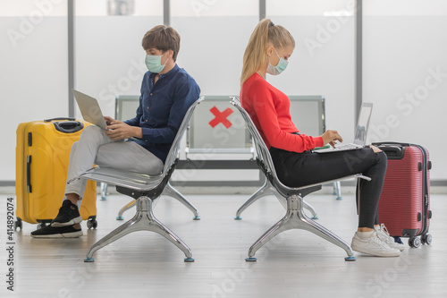 Two Caucasian couple tourists wearing hygiene protective mask on faces sitting together on chair in airport with social distancing sign. Concept for new normal traveler during coronavirus outbreak