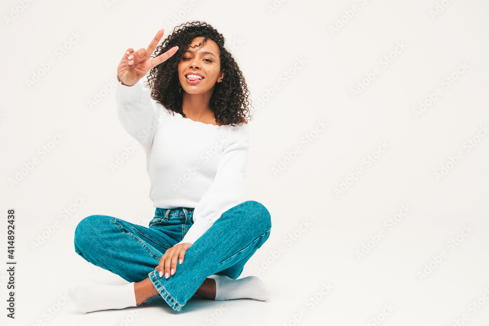 Beautiful black woman with afro curls hairstyle.Smiling model in sweater and trendy jeans clothes. Sexy carefree female sitting on white background in studio. Tanned and cheerful. Shows peace sign Beautiful black woman with afro curls hairstyle.Smiling model in sweater and trendy jeans clothes. Sexy carefree female sitting on white background in studio. Tanned and cheerful. Shows peace sign