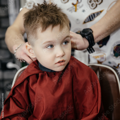 Cute little boy of five getting a haircut in a barbershop. The hairdresser does the baby's hair. Barber shop. Childhood. A new hairstyle for a little boy.