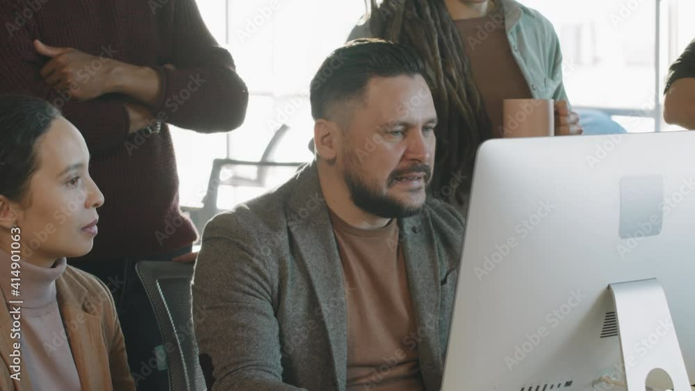 Tilt up shot of cheerful middle-aged male manager sitting at desk before computer monitor and talking to group of colleagues gathered around him Tilt up shot of cheerful middle-aged male manager sitting at desk before computer monitor and talking to group of colleagues gathered around him