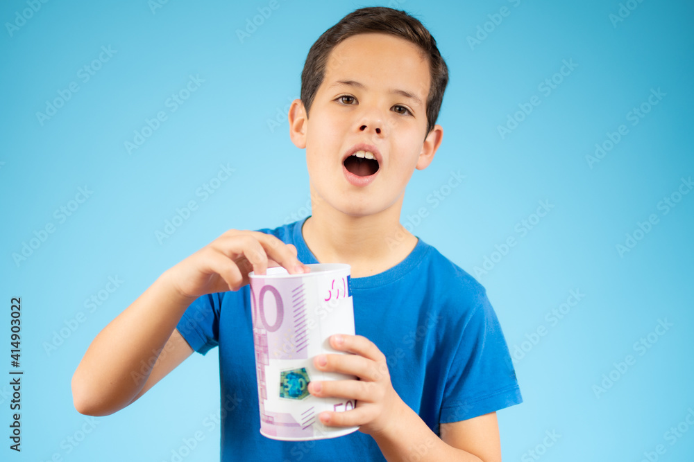 Beautiful young boy putting a coin in the piggy bank over blue background Beautiful young boy putting a coin in the piggy bank over blue background