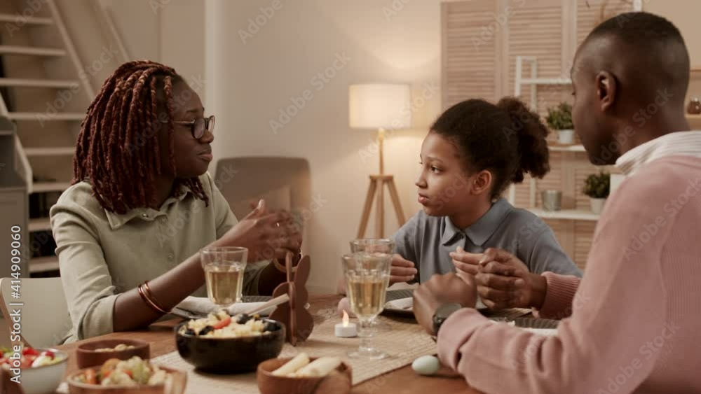 Medium close-up of school-aged African girl wearing hair up in ponytail, sitting at dinner table on festive night, looking at and listening to young mother and father having lively conversation Medium close-up of school-aged African girl wearing hair up in ponytail, sitting at dinner table on festive night, looking at and listening to young mother and father having lively conversation