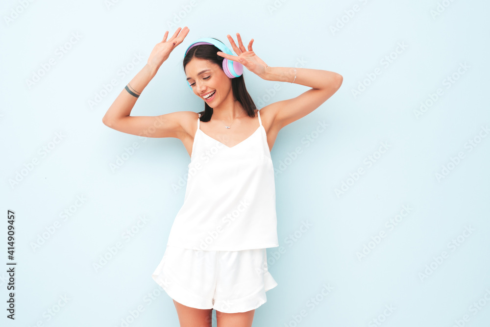 Beautiful smiling woman dressed in white pajamas. Sexy carefree model listening music in wireless headphones. Adorable and positive female posing and dancing near light blue wall in studio Beautiful smiling woman dressed in white pajamas. Sexy carefree model listening music in wireless headphones. Adorable and positive female posing and dancing near light blue wall in studio