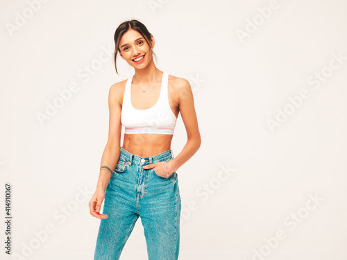 Portrait of beautiful smiling woman dressed in white jersey top shirt and jeans. Sexy carefree cheerful model having fun indoors. Adorable and positive female posing near on grey background in studio