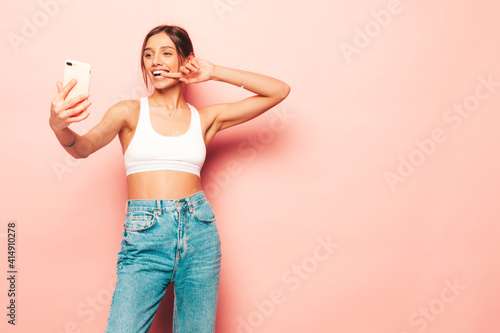 Beautiful smiling woman dressed in white jersey top shirt and jeans. Sexy carefree cheerful model having fun indoors.Adorable and positive female posing near pink wall in studio. Taking selfie