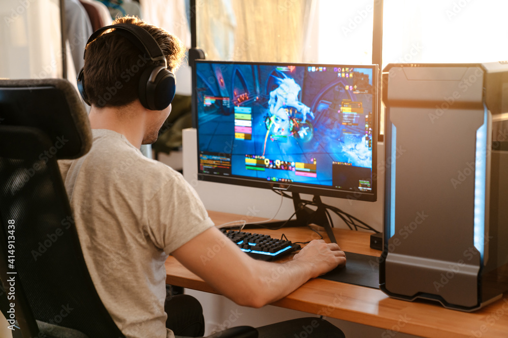 Caucasian brunette guy in headphones playing video game on his computer - obrazy, fototapety, plakaty 