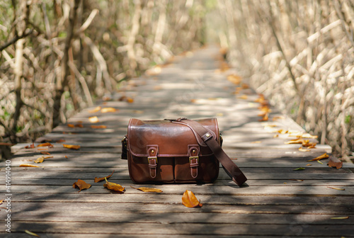 Close-up photo of dark brown leatherette bag placed on a wooden walkway. There are orange leaves around in a soft tone. Blurred backdrop of mangrove trees and bridges. Idea for traveling wallpaper. © Pang wrp