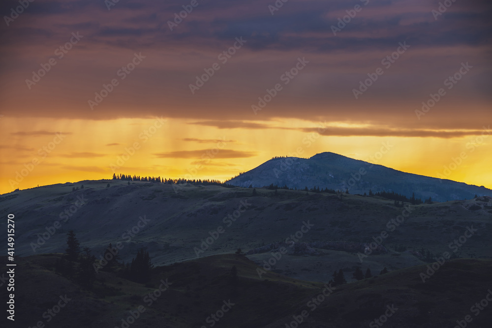 Naklejka premium Atmospheric landscape with silhouettes of mountains with trees on background of vivid orange blue lilac dawn sky. Colorful nature scenery with sunset or sunrise of illuminating color. Sundown paysage.