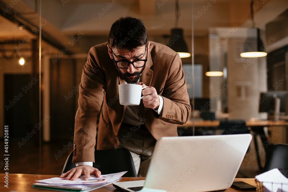 Young businessman working on laptop and drinking coffee in his office. Businessman on coffee break. Young businessman working on laptop and drinking coffee in his office. Businessman on coffee break.