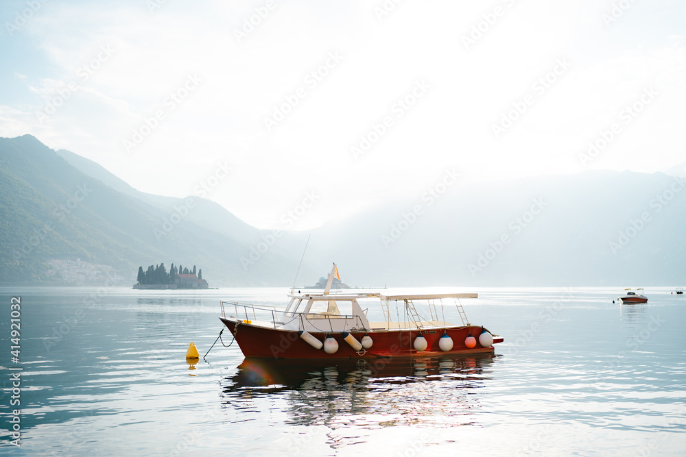 A walking red motor boat with an awning from the sun floats on the waters of the Bay of Kotor in the city of Perast against the backdrop of mountains.