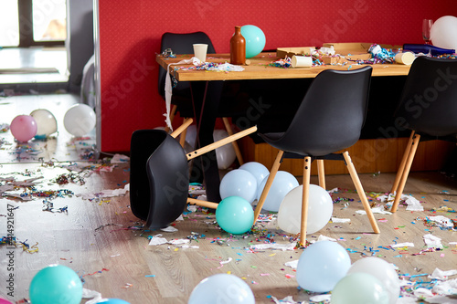 After party chaos, messy in livving room at home, table with pizza and champagne glasses covered with confetti and ballons, chair on the floor at morning after party celebration. photo