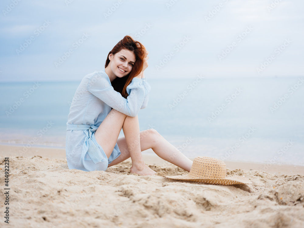 A romantic woman in a transparent blue dress sits on the sand near the sea and smiles A romantic woman in a transparent blue dress sits on the sand near the sea and smiles