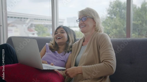Medium shot - Adopted Asian daughter and her Caucasian stepmother enjoy watching movie on internet at home. Love and caring time of Multi-racial family concept.