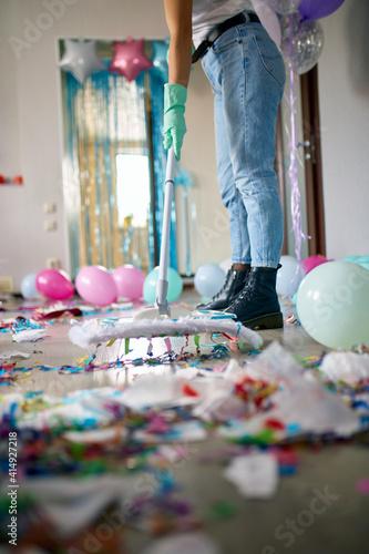 Fotografie Woman with pushbroom cleaning mess of floor in room after party confetti