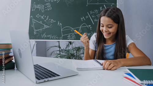 happy girl holding pen and looking at notepad near laptop on desk