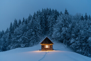 Fantastic winter landscape with glowing wooden cabin in snowy forest. Cozy house in Carpathian mountains. Christmas holiday concept