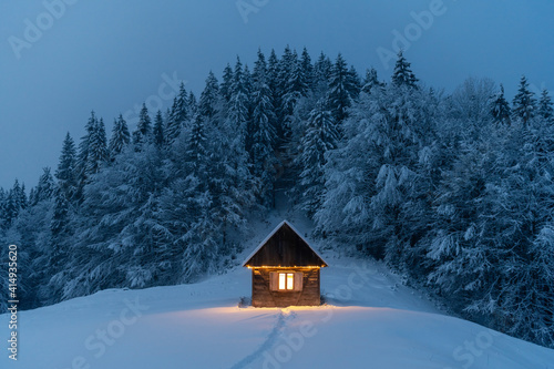 Fantastic winter landscape with glowing wooden cabin in snowy forest Fototapeta