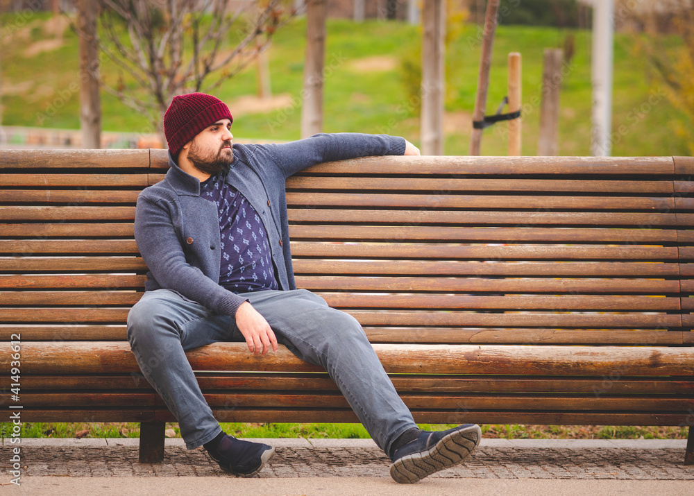 Boarded guy in a woolen hat and sweater sitting on park bench relaxing. Boarded guy in a woolen hat and sweater sitting on park bench relaxing.
