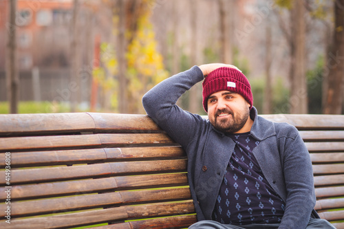 Boarded guy in a woolen hat and sweater smiling and sitting on park bench.