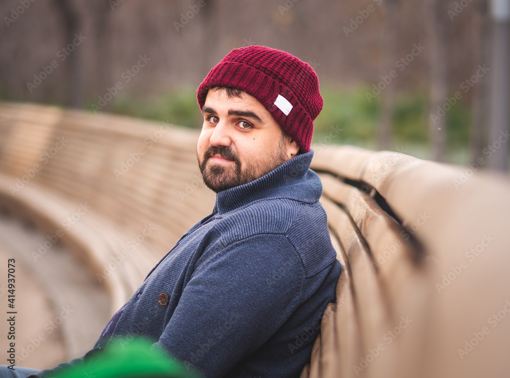 Boarded guy in a woolen hat and sweater sitting on park bench and looking at the camera. Boarded guy in a woolen hat and sweater sitting on park bench and looking at the camera.