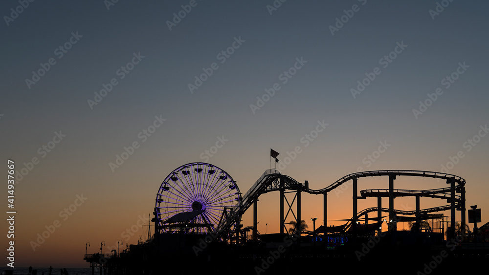 Silhouette of the popular ferris wheel known as the Pacific Wheel and the steel roller coaster known as The Santa Monica West Coaster, on the popular pier at the sunset in Los Angeles, California, USA