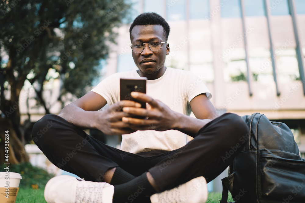 Fototapeta premium Pensive dark skinned male student in eyewear for vision correction sitting outdoors on street with backpack and mobile phone, serious african american hipster guy blogger checking mails on smartphone