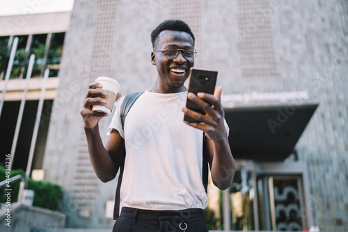 Happy dark skinned male millennial in casual wear enjoying watching videos from vlogs on smartphone outdoors  smiling african american man holding coffee cup and searding good news on mobile phone