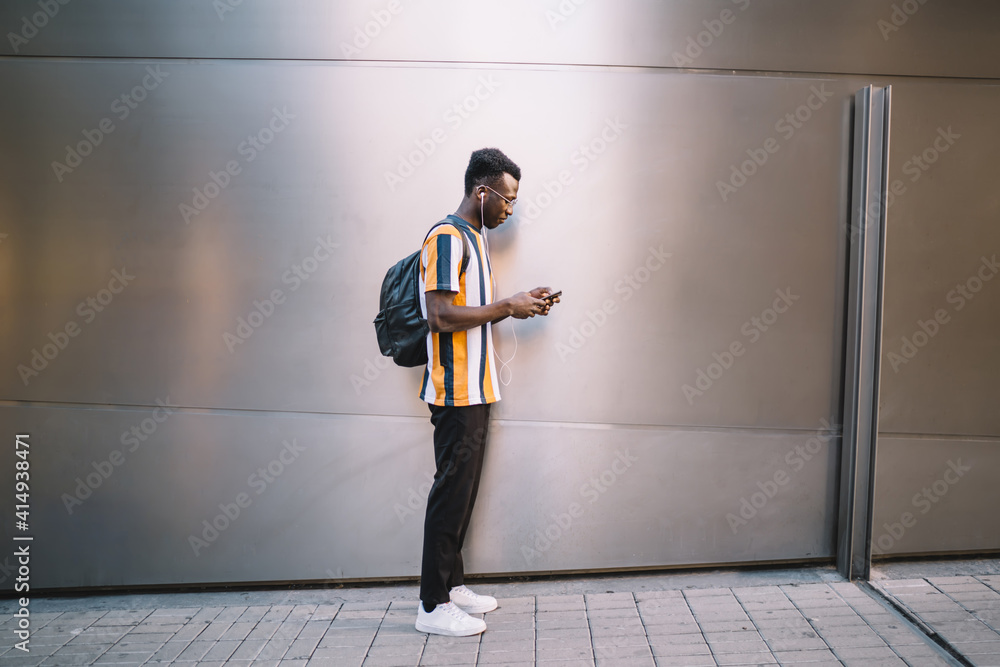 Dark skinned male blogger connecting to 4g wireless internet for browsing website with content text, millennial hipster guy in earphones choosing music playlist for listen during travel walk in city Dark skinned male blogger connecting to 4g wireless internet for browsing website with content text, millennial hipster guy in earphones choosing music playlist for listen during travel walk in city