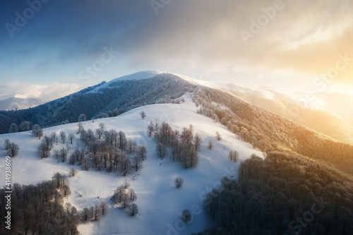Amazing aerial view of mountains range  meadows and snow-capped peaks in winter time. Forest with frost glowing with bright warm sunrise light