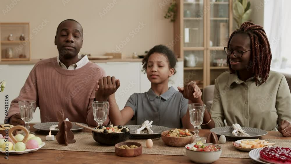 Medium close-up of happy African man talking to school-aged daughter, smiling, then girl holding hands with mother and father, family sitting at festive dinner table, closing eyes and praying Medium close-up of happy African man talking to school-aged daughter, smiling, then girl holding hands with mother and father, family sitting at festive dinner table, closing eyes and praying