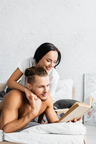 Smiling woman embracing shirtless boyfriend with book on bed