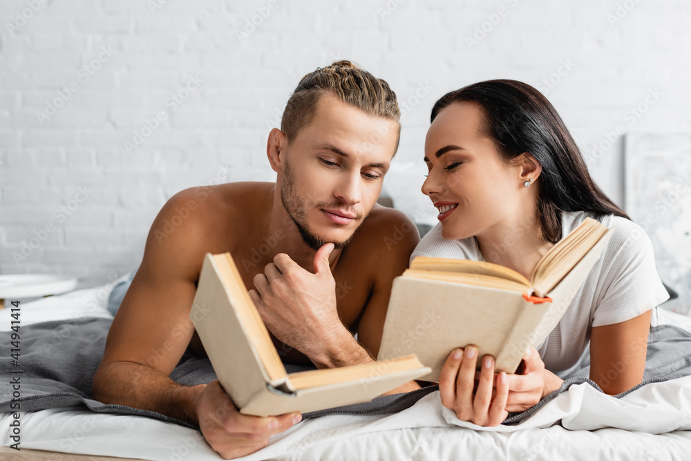 Smiling woman holding book and looking at shirtless boyfriend on bed Smiling woman holding book and looking at shirtless boyfriend on bed