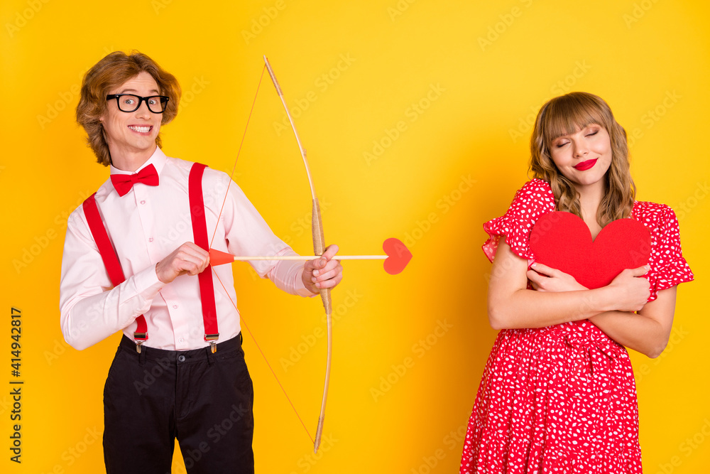Photo portrait of guy with arrow bow and funky girl fell in love embracing red heart isolated on vivid yellow background Photo portrait of guy with arrow bow and funky girl fell in love embracing red heart isolated on vivid yellow background