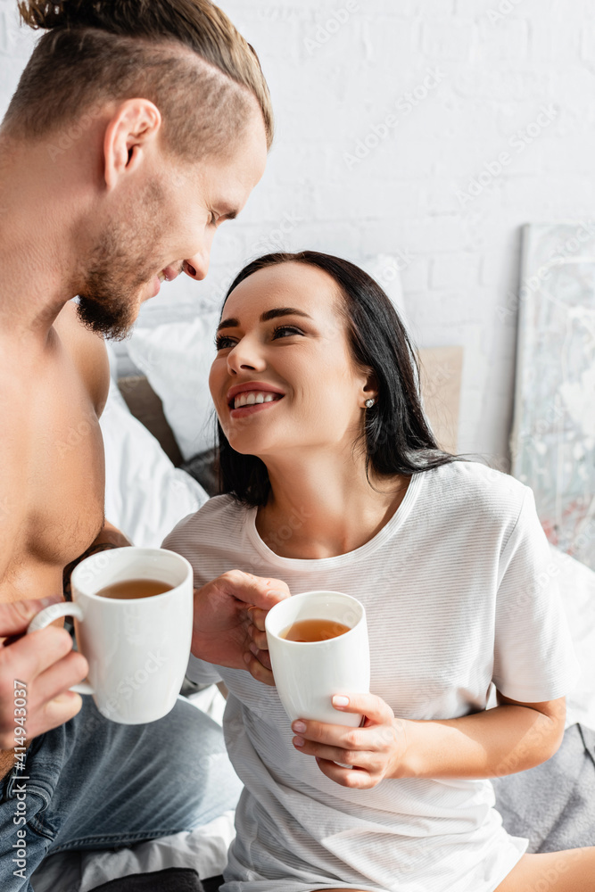 Smiling woman looking at sexy man with cups of tea on bed Smiling woman looking at sexy man with cups of tea on bed
