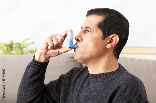 Side view of an asthmatic man using an inhaler sitting on a couch in the living room at home