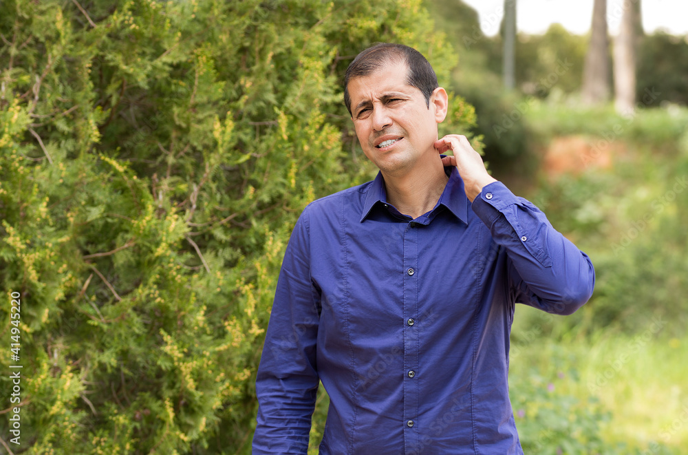 Man suffering itching scratching neck standing outdoors in a park Man suffering itching scratching neck standing outdoors in a park
