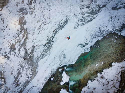 Drone shot of hiker with backpack, orange cap and orange pants hiking on dangerous path through snow and ice next to cold mountain river in austria