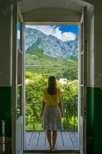 Inside a dark room with a woman looking into the distance through the wide-open doors that reveal the top of a mountain on a sunny summer day, a contrasting image that gives the idea of ​​hope.