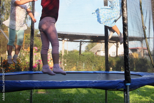 A three little Russian girls jumps on a outdoor 8ft trampoline with outside safety soft net fence on green grass in the rural Garden at Sunny summer day, jumping kids active sport vacation photo